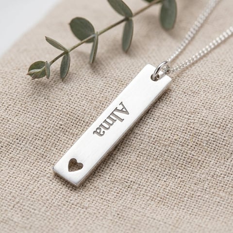 Silver rectangular pendant necklace engraved with BLUNT text and small heart cutout, on beige linen with dried eucalyptus leaves