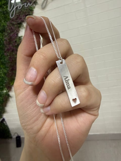 Hand holding a silver nameplate necklace with Alicia engraved on it, featuring a small heart cutout